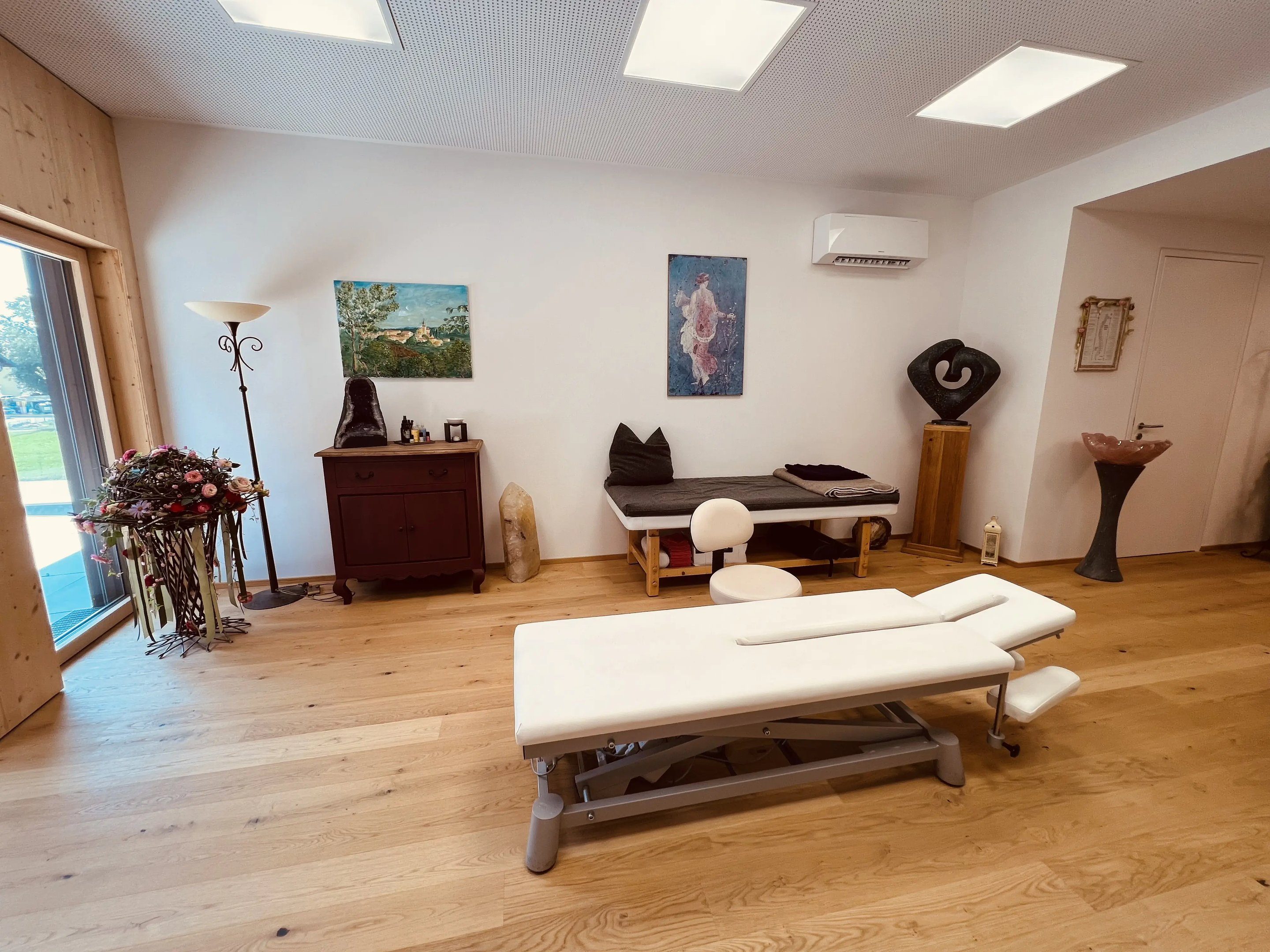 Heller Praxisraum der Atlaspraxis Seeham mit Liege, Holzboden und Glastür zum Garten